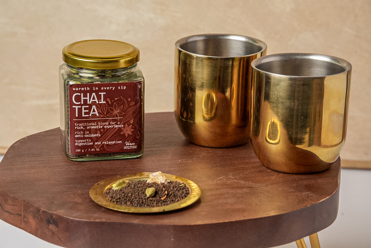 Chai tea spice blend in glass jar with gold lid displayed with two brass cups and spices on wooden tray.