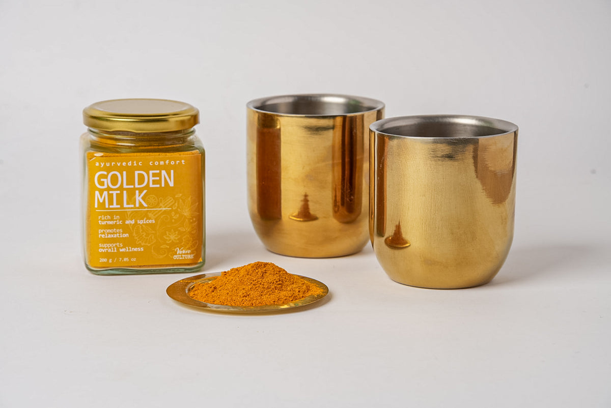 Golden milk spice blend in glass jar with white label, displayed with two gold metal cups and powdered spice on spoon.