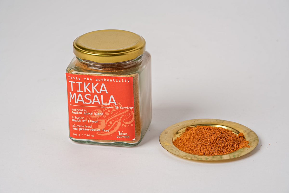 Glass jar with gold lid containing reddish-brown tikka masala spice blend, with a small wooden dish of spice beside it.