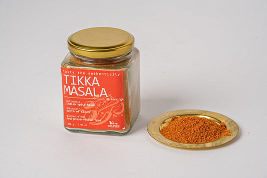 Glass jar with gold lid containing reddish-brown tikka masala spice blend, with a small wooden dish of spice beside it.