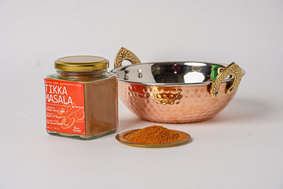 Glass jar of tikka masala spice blend with red label, wooden spoon with spice powder, and copper serving bowl with brass hand