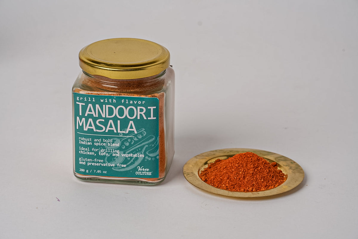 Glass jar with gold lid containing reddish-brown tandoori masala spice blend, with wooden dish displaying the spice powder be