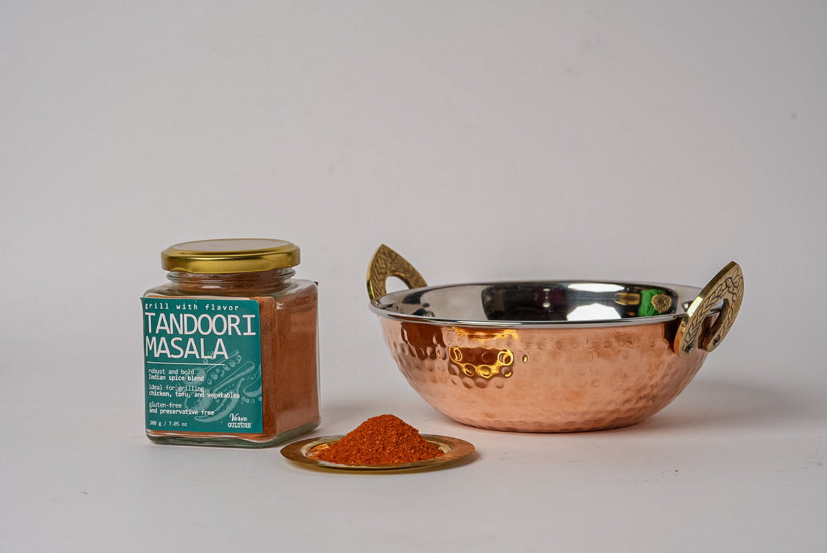 Tandoori masala spice blend in glass jar with gold lid, alongside small brass dish of reddish-brown powder and copper serving