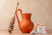 Wooden molinillo whisk, terracotta pitcher, and cloth pouch containing hot chocolate mix on neutral background.