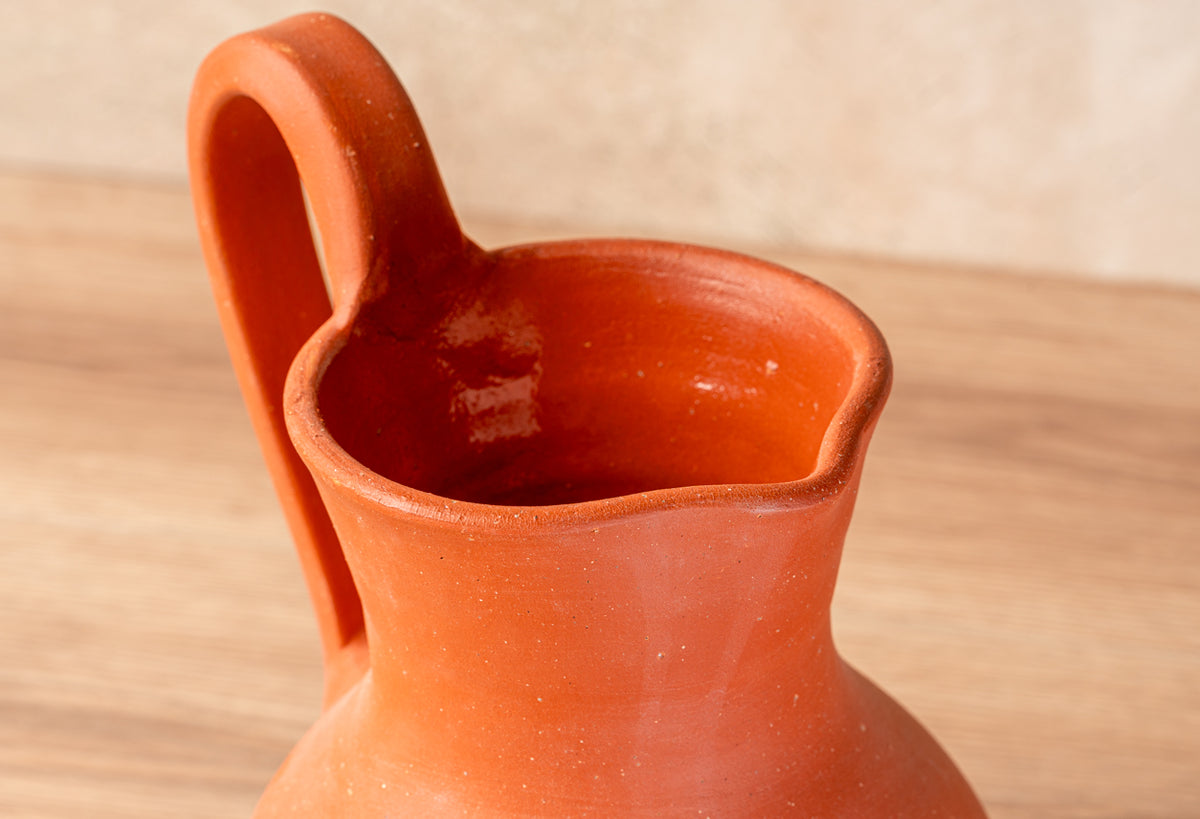 Red-orange ceramic jug with wide handle and rounded body on wooden surface.