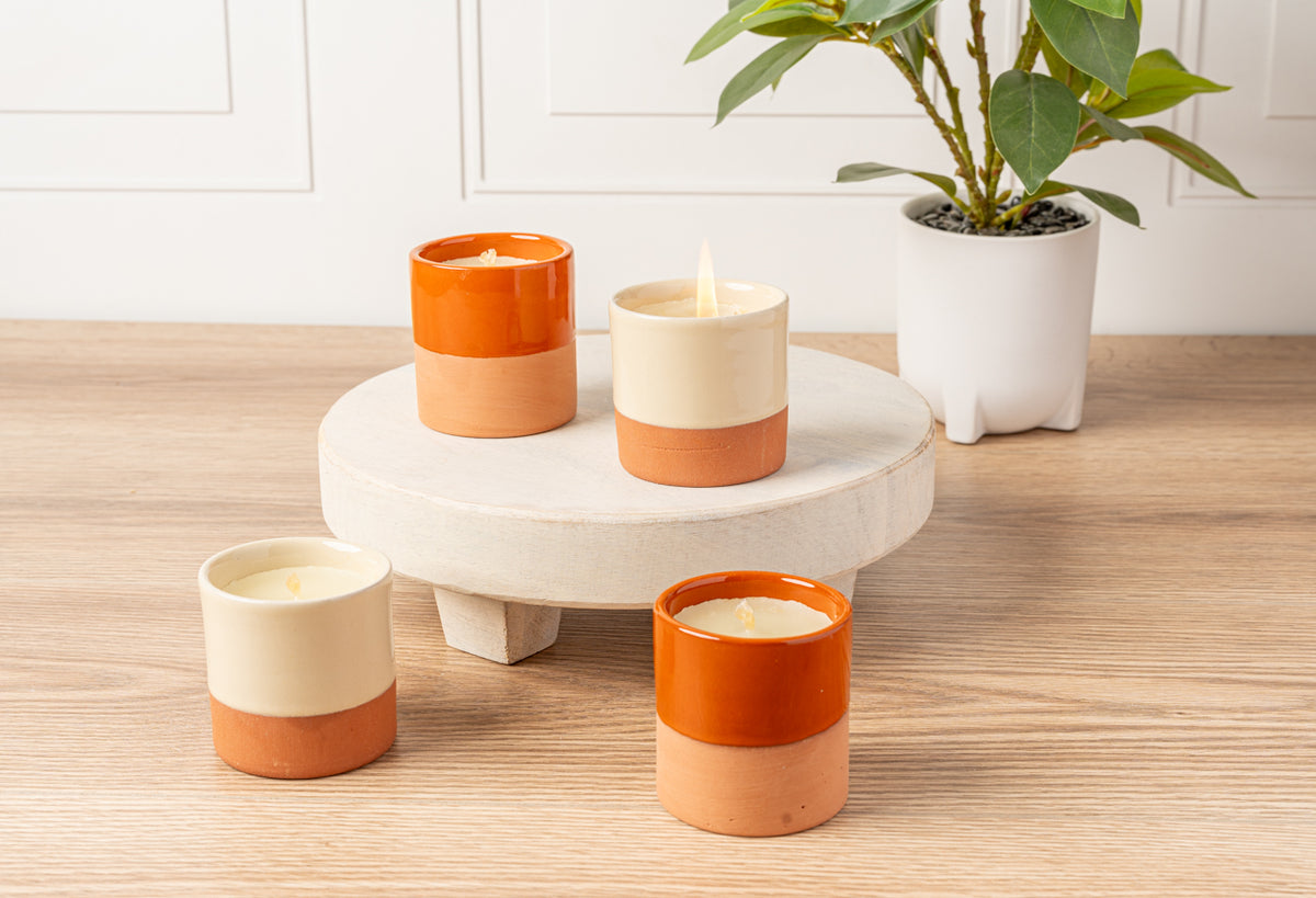 Four terracotta and cream cylindrical soy candles with lit flames displayed on a white pedestal and wooden surface.