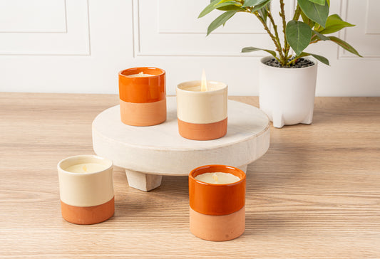 Four terracotta and cream cylindrical soy candles with lit flames displayed on a white pedestal and wooden surface.