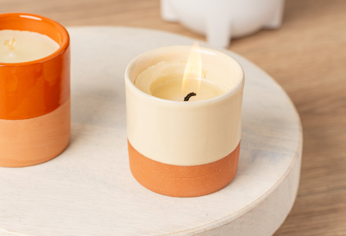 Lit candle in cream and terracotta ceramic vessel with matching unlit candle on wooden surface.