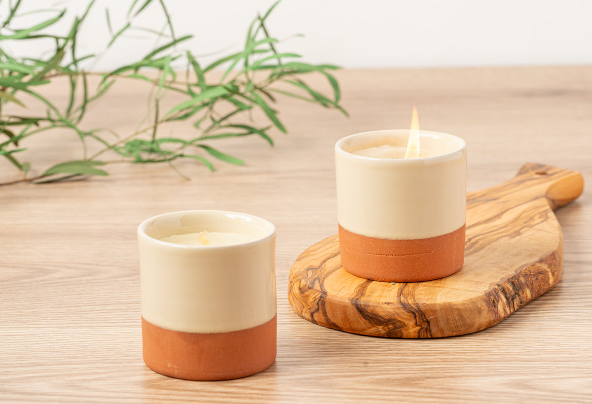 Two cream and terracotta soy candles with lit wicks on wooden coasters with greenery in background.