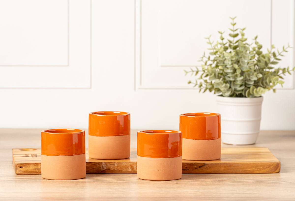 Four terracotta cups with orange glazed tops and natural clay bases arranged on a wooden board.
