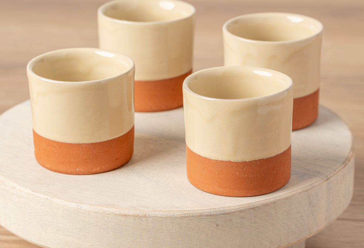 Four cylindrical cups with cream-colored glaze on top and terracotta base arranged on a round wooden platform.