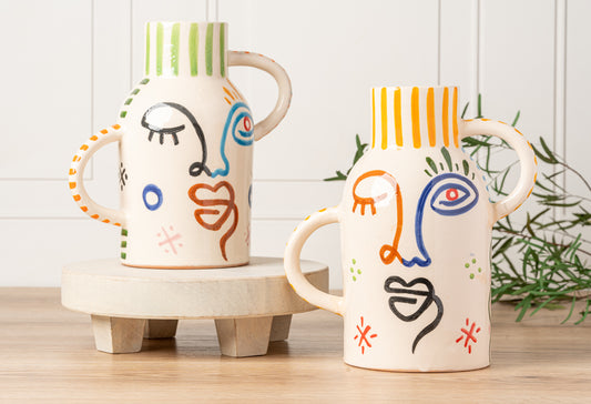 Two cream ceramic vases with abstract hand-painted faces, striped rims, and natural handles in green, blue, orange, and black