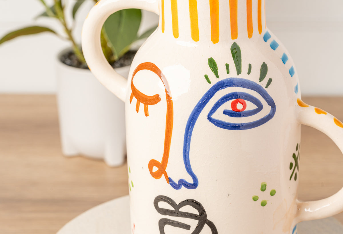 Cream ceramic vase with hand-painted abstract face featuring blue eye, orange and green details, and curved handles.