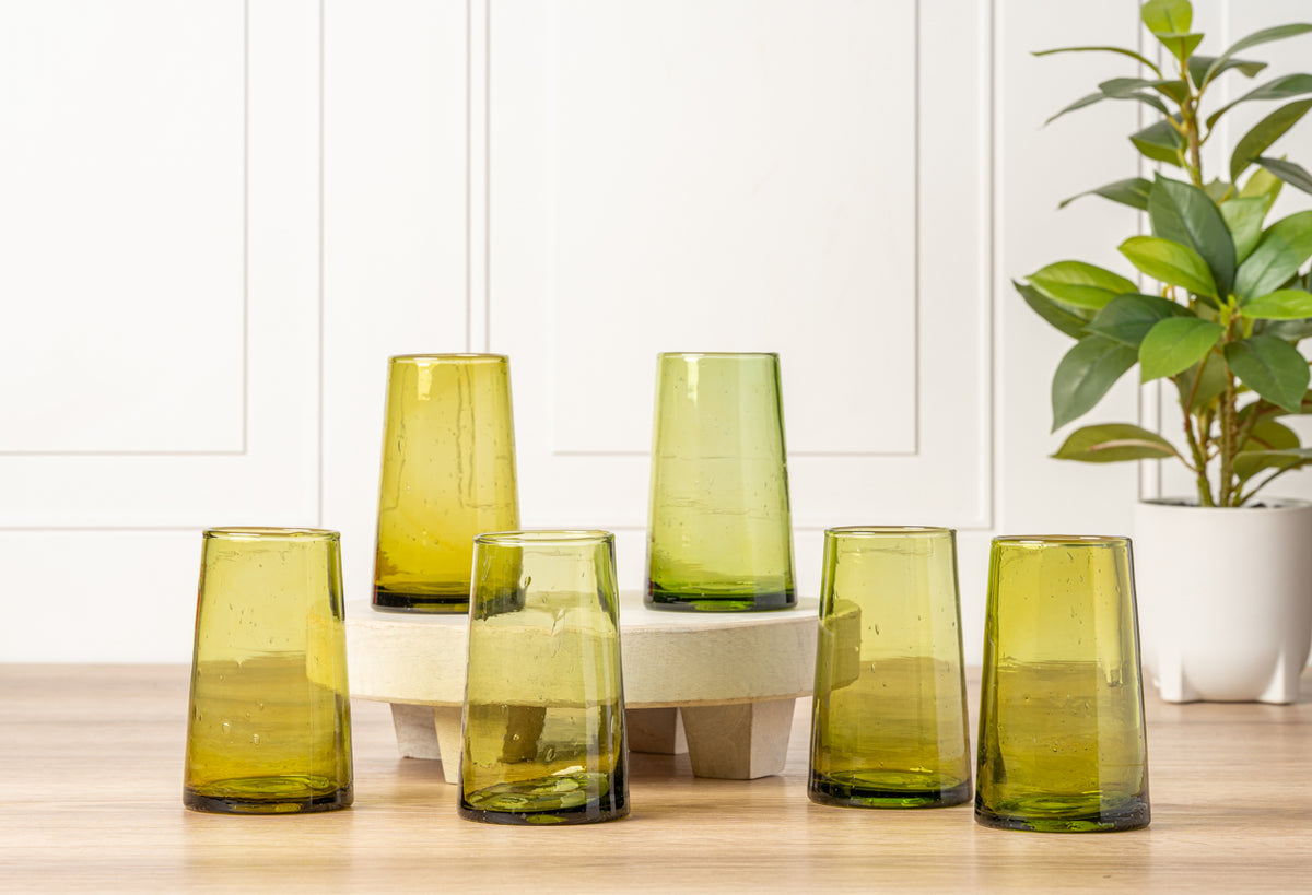 Six cone-shaped glasses in yellow and green tones arranged on a wooden surface with a potted plant.