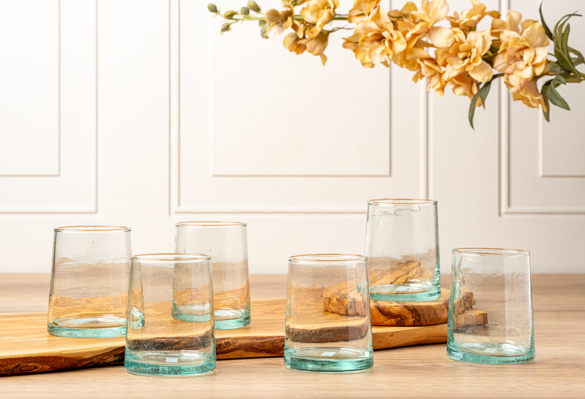 Six clear glasses with gold rim and turquoise base stripe in varying heights on wooden surface.