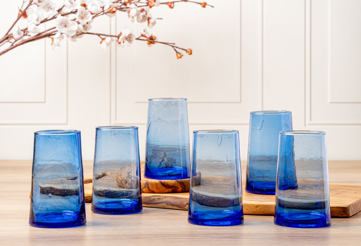 Six blue cone-shaped glasses with layered blue and amber glass, displayed on wood surface with dried branches.