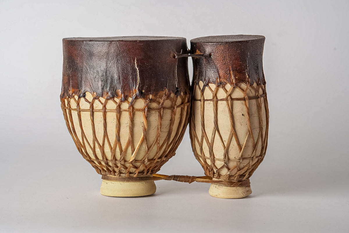 Two connected goblet-shaped drums with dark leather tops, cream-colored wooden bodies, and woven rope tuning around the sides