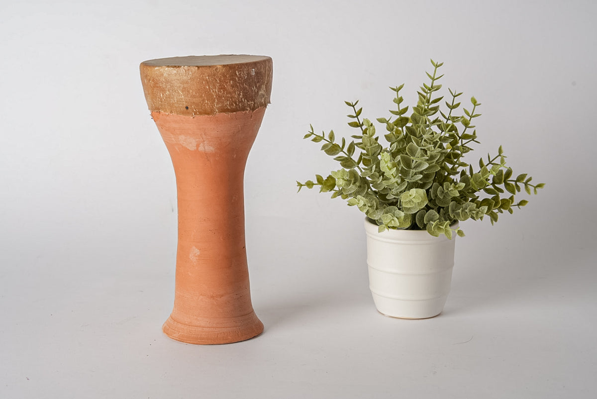Terracotta goblet drum with textured brown top and cylindrical stem, displayed next to potted green plant.