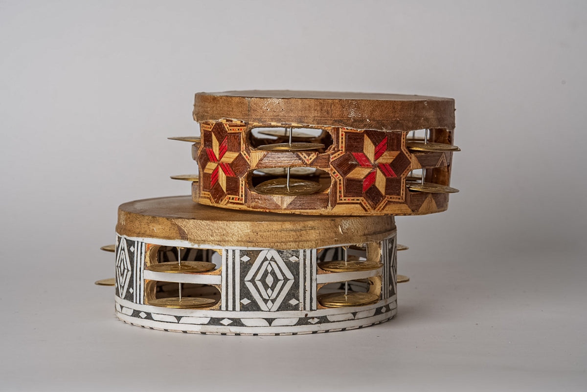 Two wooden frame drums with metal jingles, decorated with geometric patterns in red and white inlay, stacked together.