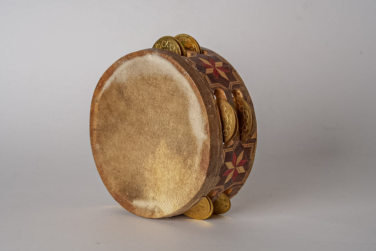 Round wooden tambourine with cream-colored drumhead, brass jingles, and decorative inlaid patterns on the wooden frame.
