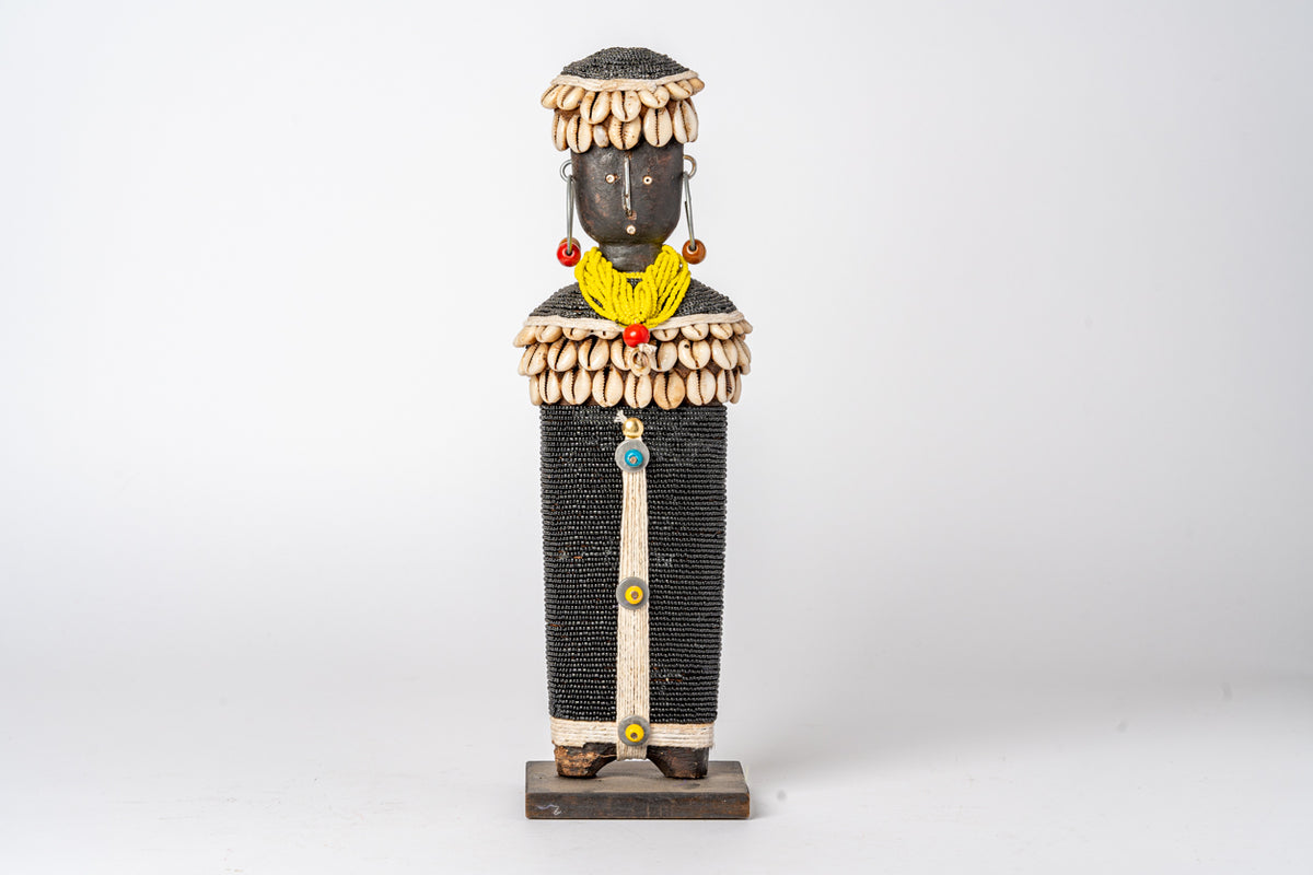Carved wooden doll with black beaded body, cowrie shell necklace and headdress, yellow wrapped neck detail, and red bead earr