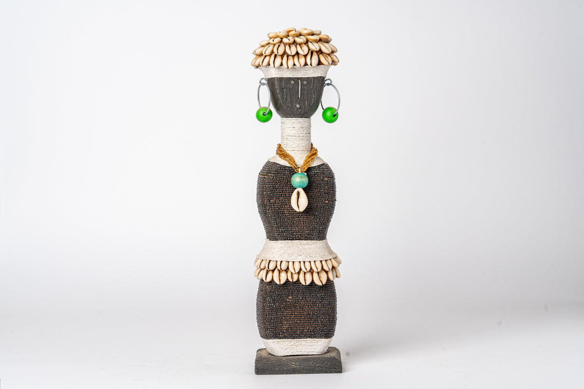 Stacked decorative doll with woven charcoal sections, shell and bead details, green earrings, and cowrie shell pendant.