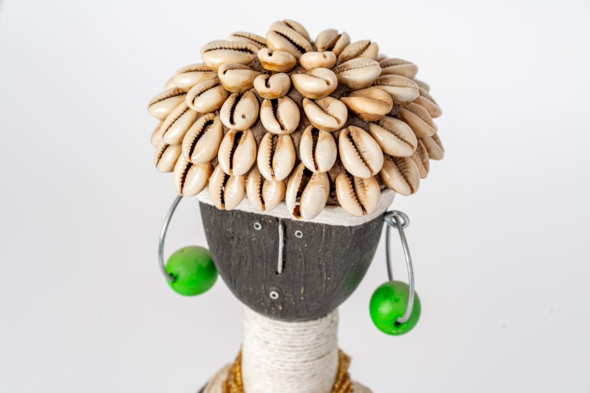 Decorative doll with cowrie shell hair, dark wooden face, wrapped stem neck, and two green bead earrings.