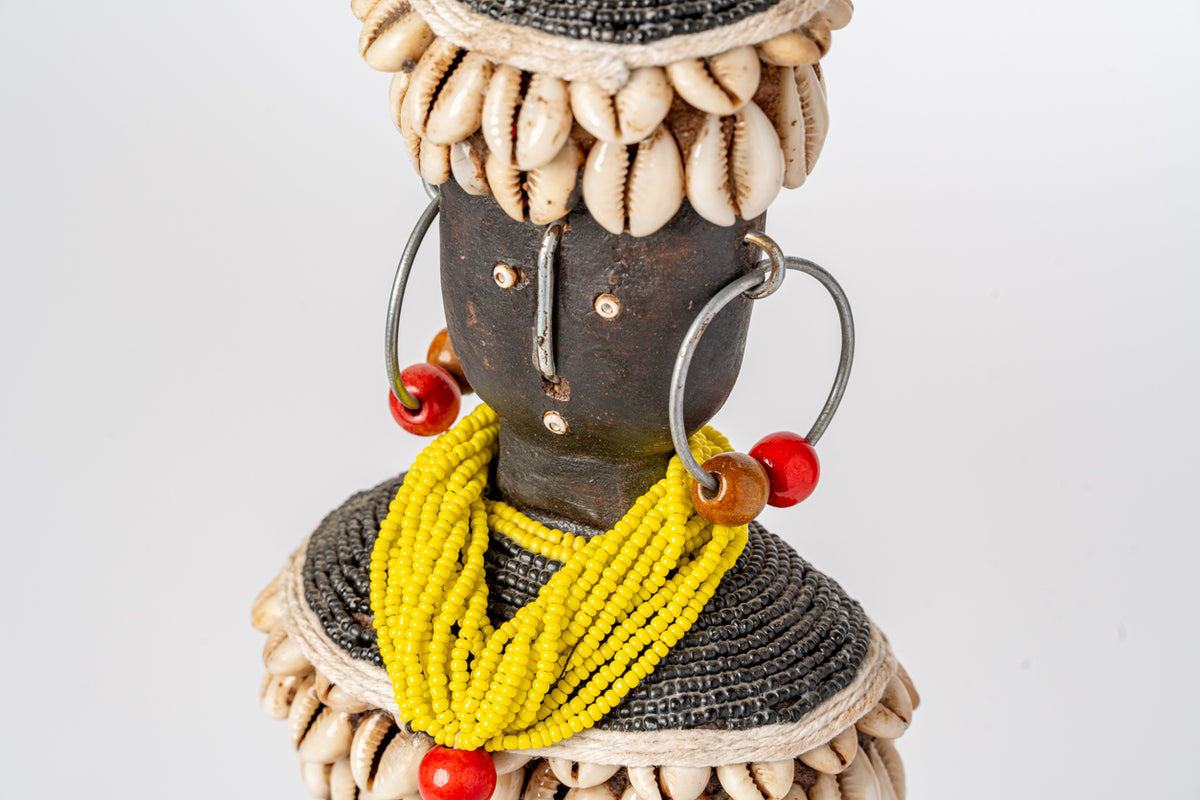 Wooden doll with dark carved head, cowrie shell collar, yellow beaded necklace, and red and orange beads.