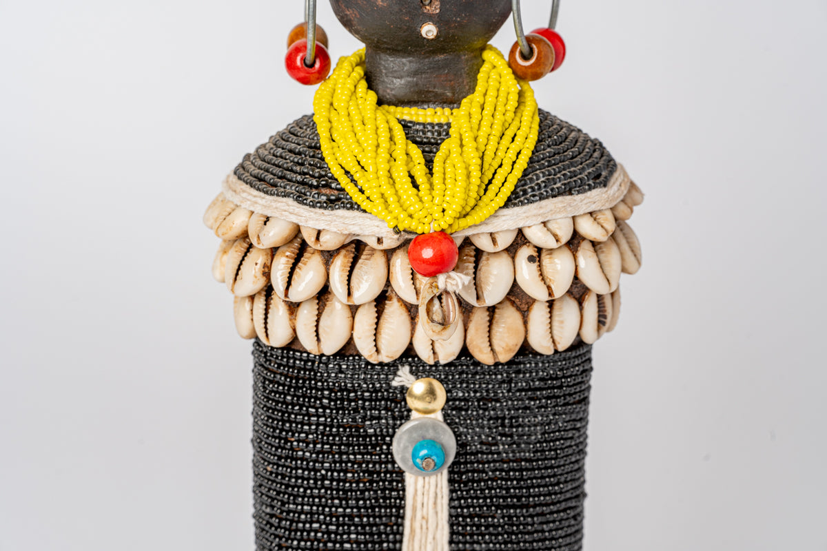 Wooden doll with dark carved head, yellow beaded necklace, shell chest piece, red and orange beaded earrings, and black beade