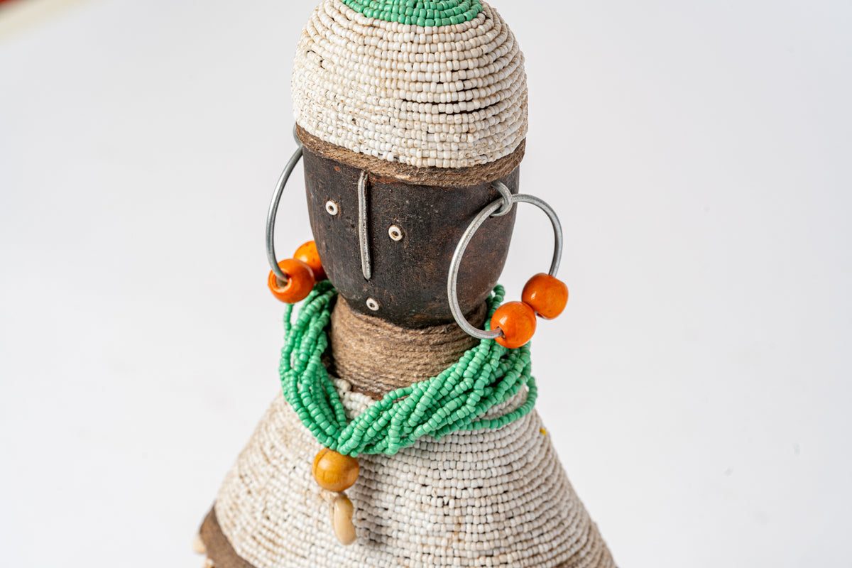 Beaded doll with woven cream and green body, dark face, wire arms with orange beads, and green beaded necklace with orange ac