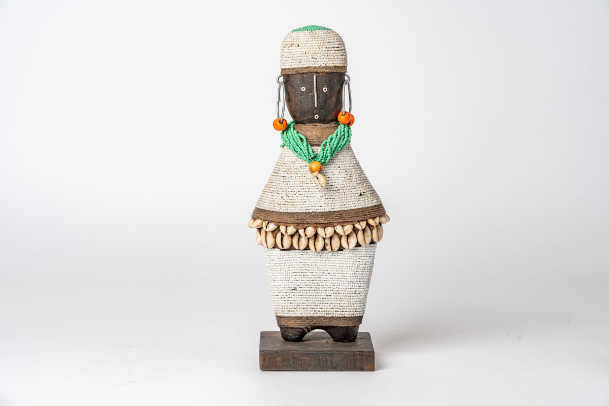 Wooden doll with wrapped beige fiber body, dark wood head and base, green beaded necklace, and orange bead accents.