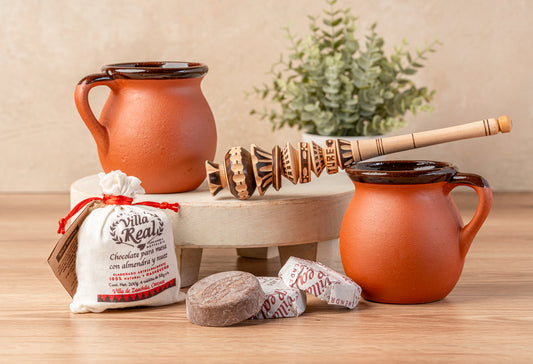 Two terracotta pitchers, wooden whisk, chocolate tablets, and packaged mix arranged on a white ceramic tray.