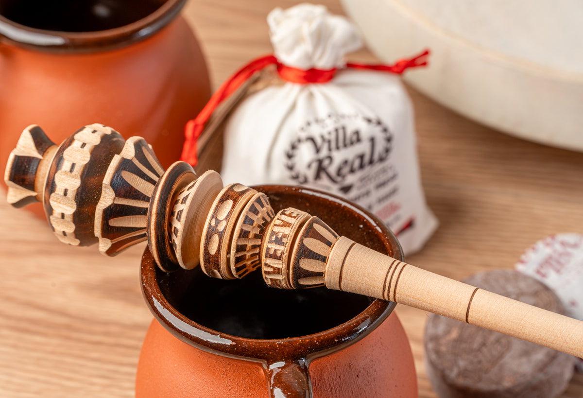Decorative wooden molinillo whisks resting in a clay pot with an orange exterior, accompanied by a cloth gift bag on a wooden