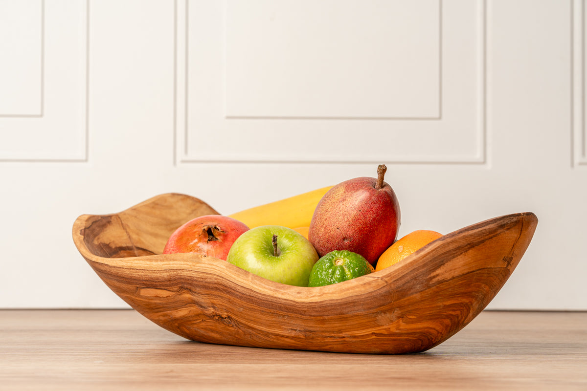 Wooden bowl with natural grain and irregular edges holding apples, pear, and citrus fruits on white paneled background.