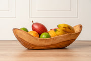 Wooden boat-shaped bowl filled with apples, bananas, oranges, and limes on a wooden surface.