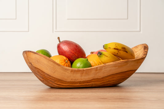 Wooden boat-shaped bowl filled with apples, bananas, oranges, and limes on a wooden surface.