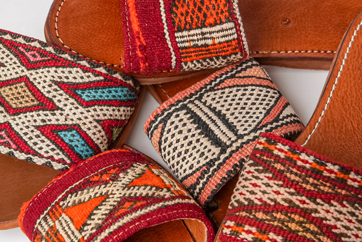 Tan leather slides with woven kilim textile uppers in red, orange, cream, and blue geometric patterns.