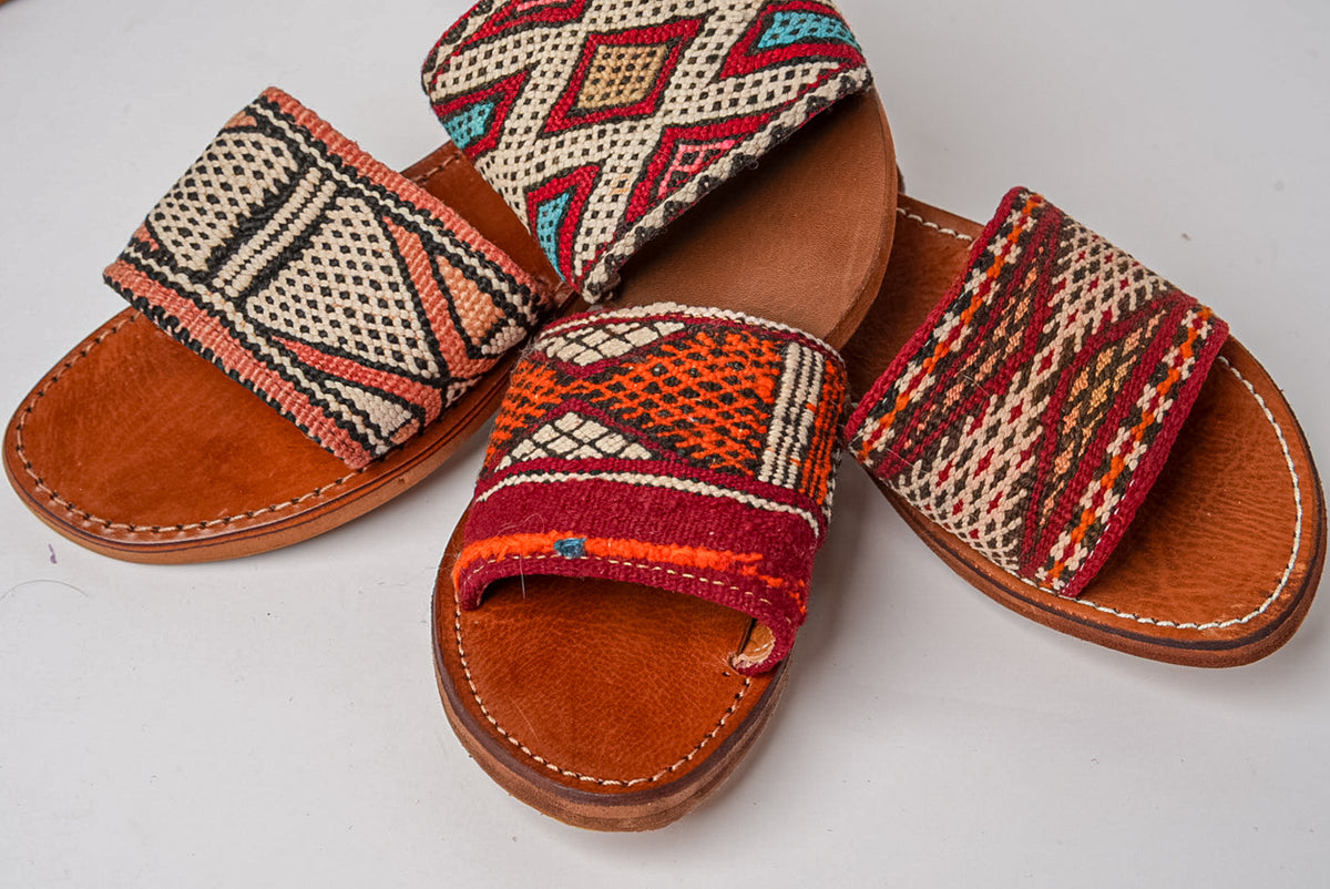 Three tan leather slides with geometric patterned fabric uppers in red, cream, blue, and orange tones.