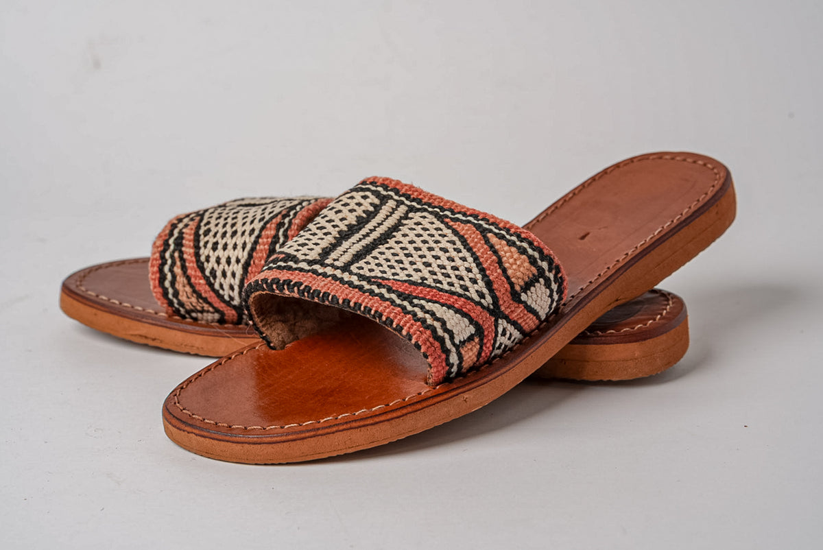 Flat slide sandals with tan leather sole and woven geometric patterned upper in cream, black, and rust tones.