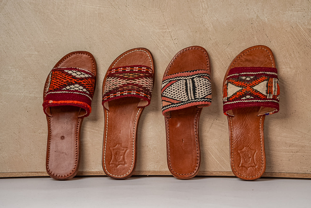 Four brown leather slides with geometric patterned fabric straps in red, cream, and black.