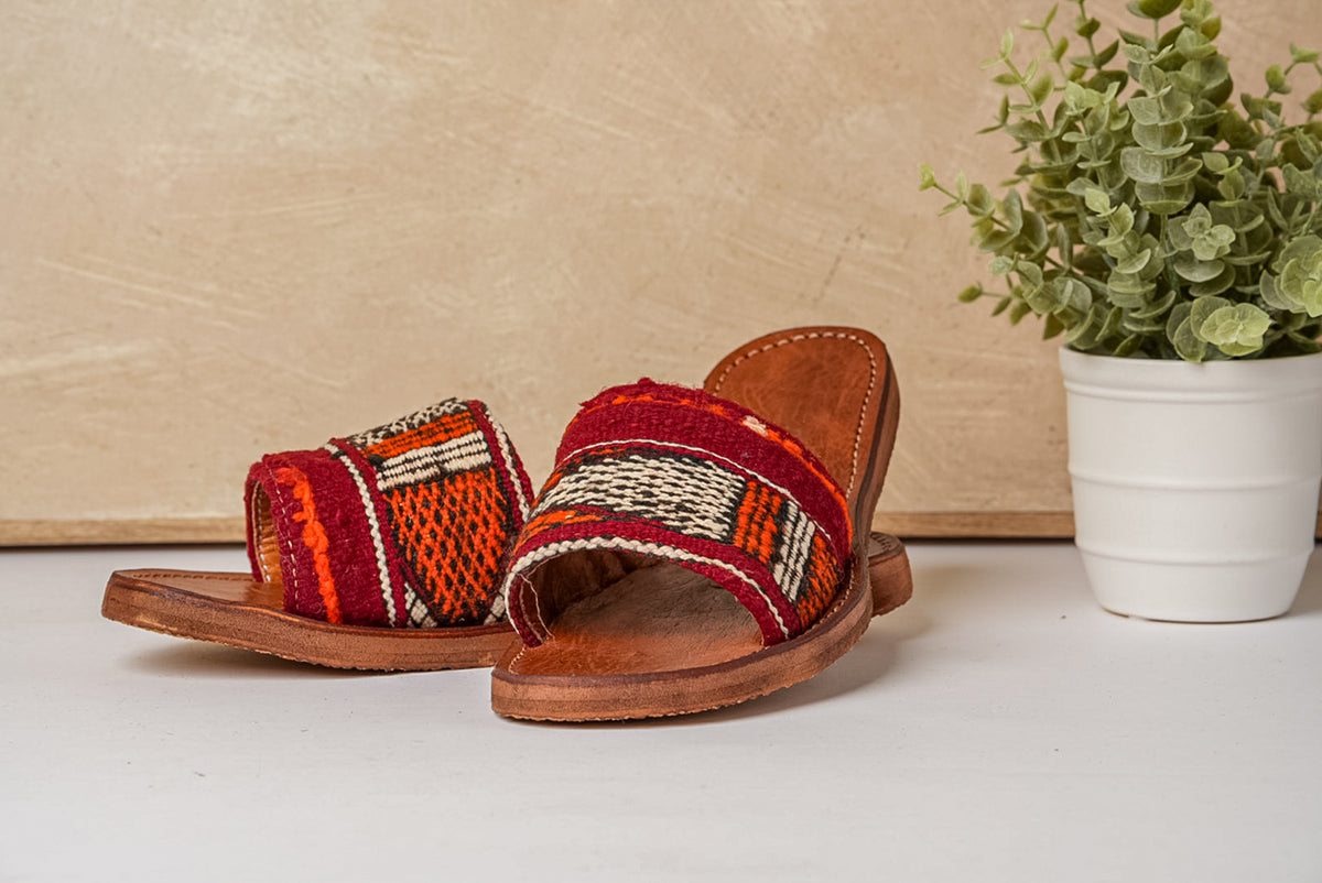 Pair of slide sandals with red and orange woven textile uppers and brown leather soles.