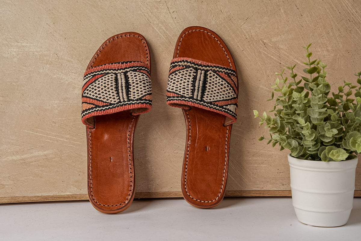 Pair of brown leather slides with woven geometric patterned straps in cream and black.