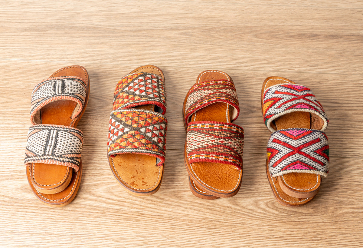 Four tan leather slides with woven textile straps in geometric patterns displayed on a wooden surface.