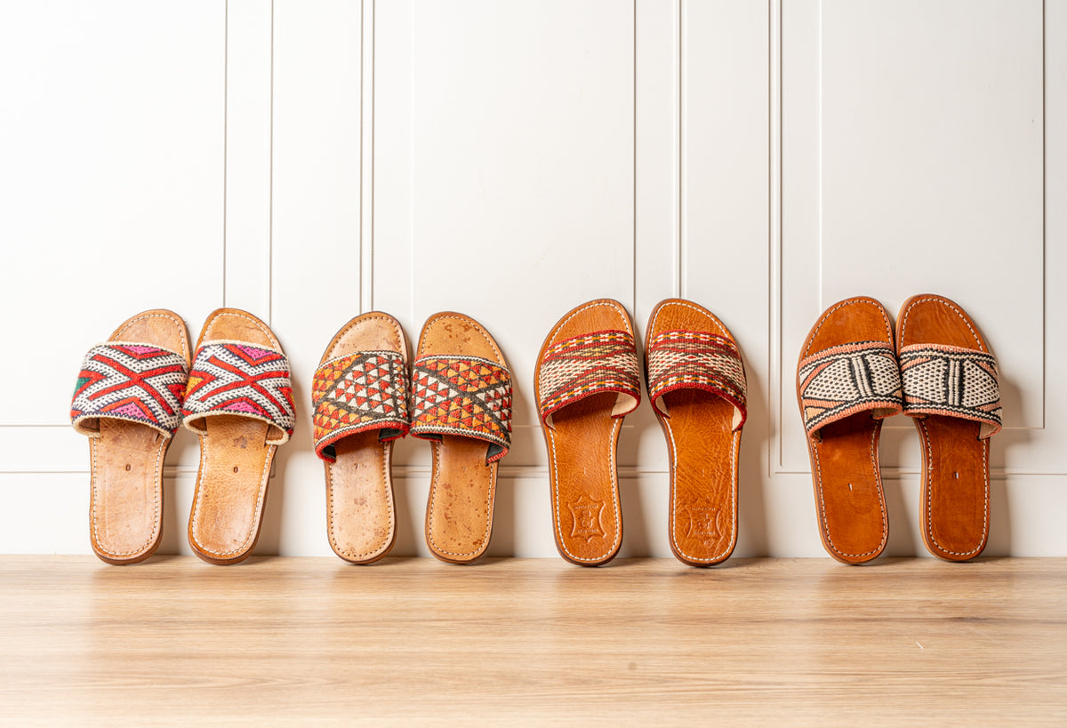 Four pairs of cork slides with woven textile uppers in varying geometric patterns and orange tones.