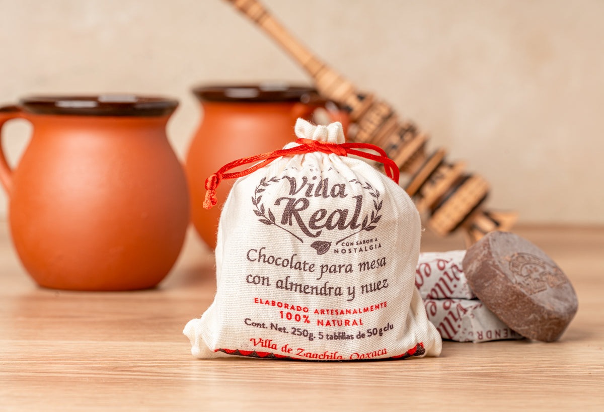 Muslin bag with "Villa Real" chocolate mix label, two orange ceramic cups, wooden whisk, and chocolate tablet on wooden surfa