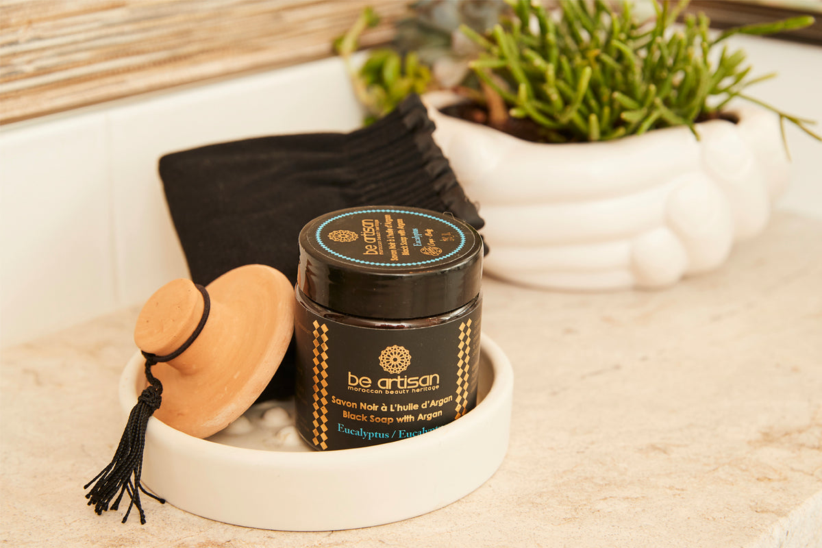Black soap jar with argan oil, wooden exfoliating disc with tassel, and black washcloth displayed on marble surface with succ