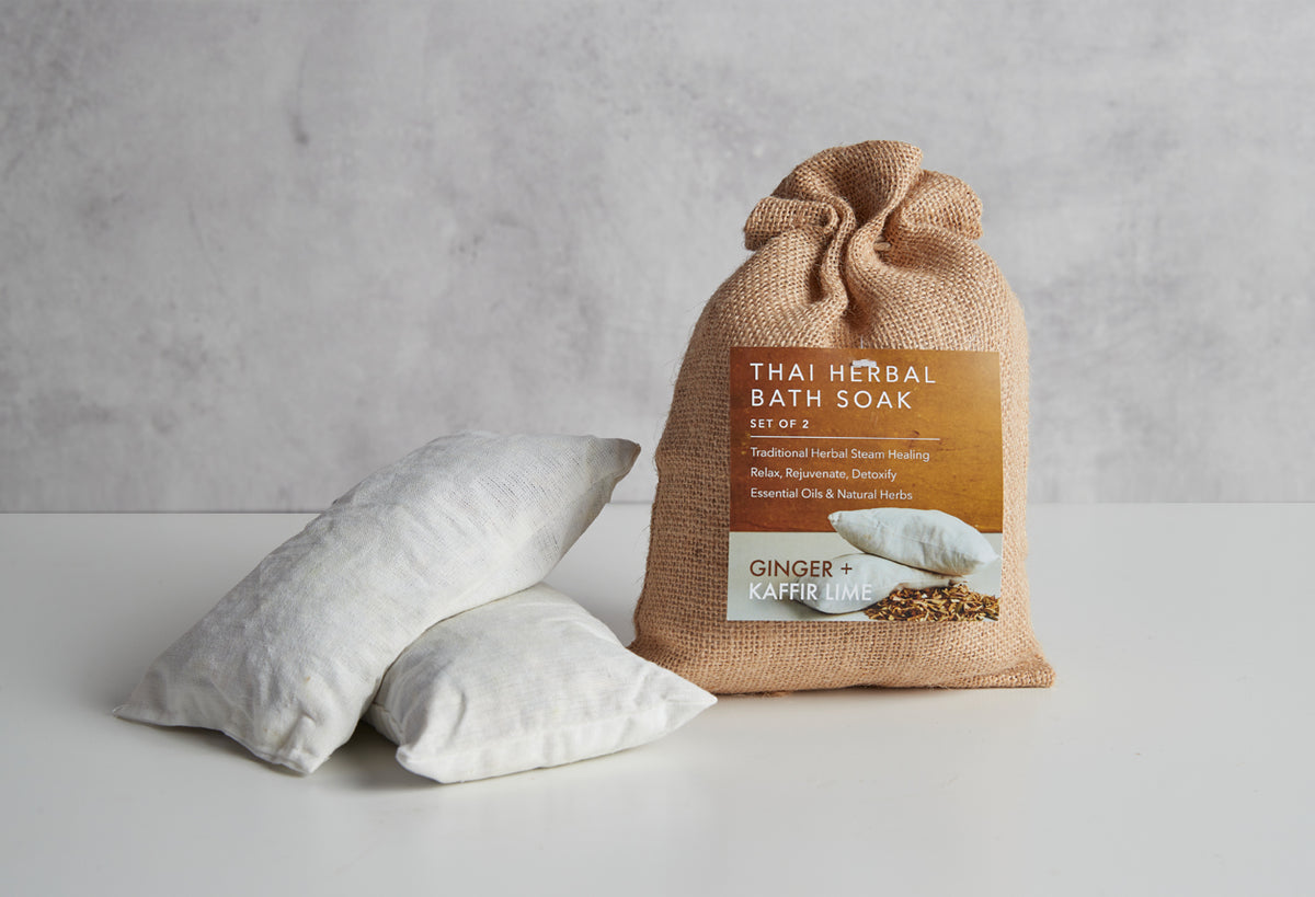 Tan burlap bag with brown and cream label next to two white muslin bath soak pillows on white surface.