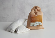 Tan burlap bag with brown and cream label next to two white muslin bath soak pillows on white surface.