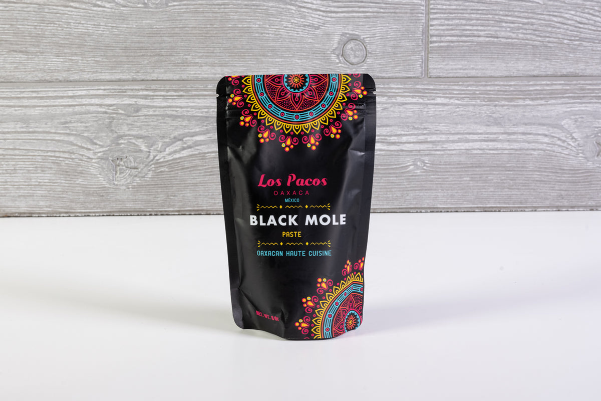 Black pouch labeled Los Pacos Black Mole Paste with colorful geometric pattern design on white wood background.