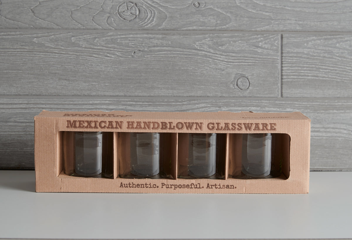 Set of four hand-blown Mexican glassware cups in cardboard packaging against wooden background.